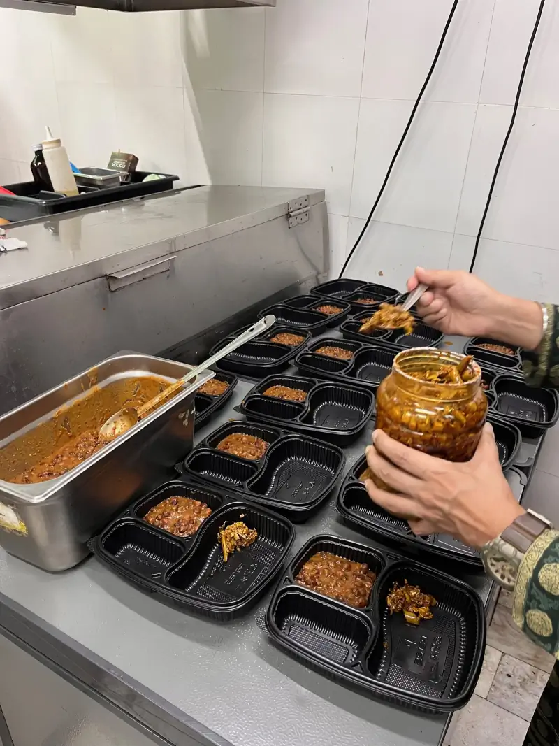 A jar of achaar being spooned into each meal tray beside a tray of hot chhole