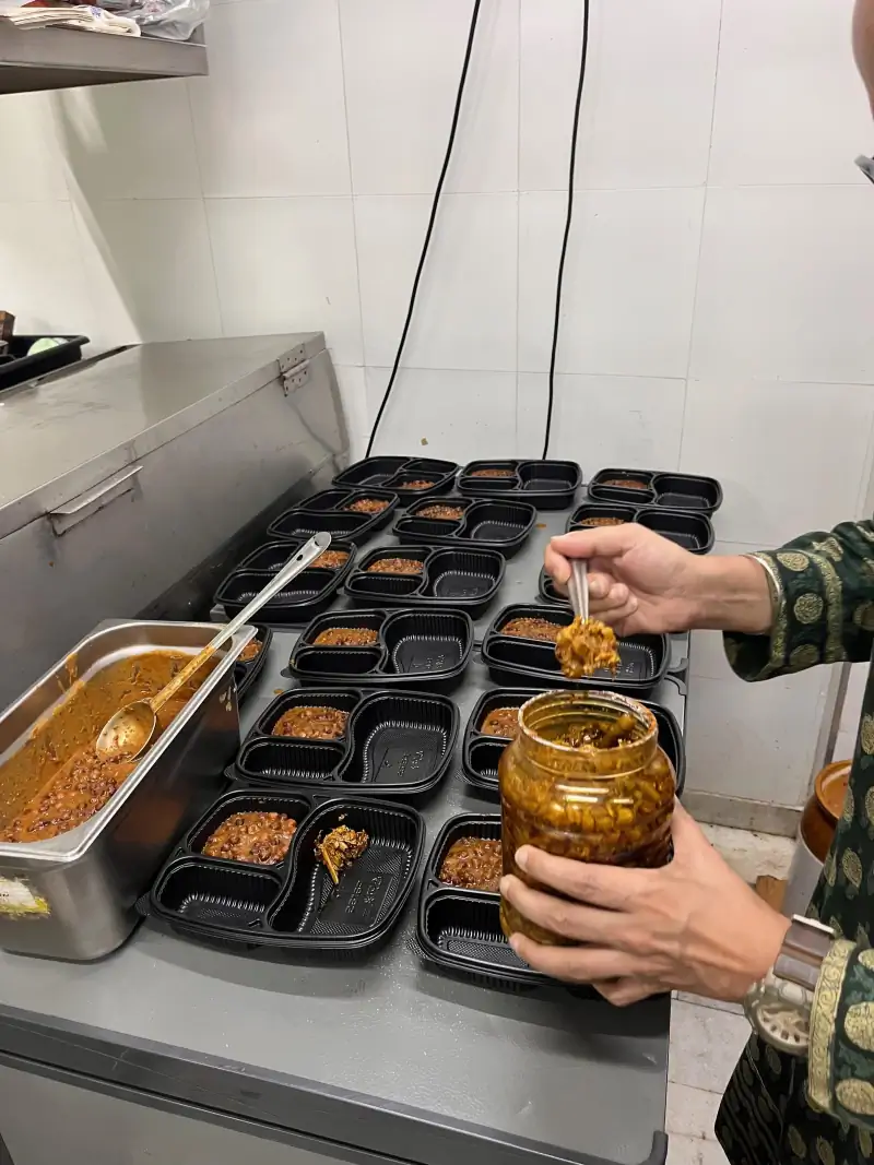 Hands spooning chhole into sectioned meal trays laid out across a steel counter